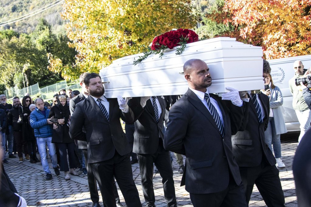Pamela Genini, a model, wearing sunglasses and a patterned dress, sitting with a small white dog and a handbag.